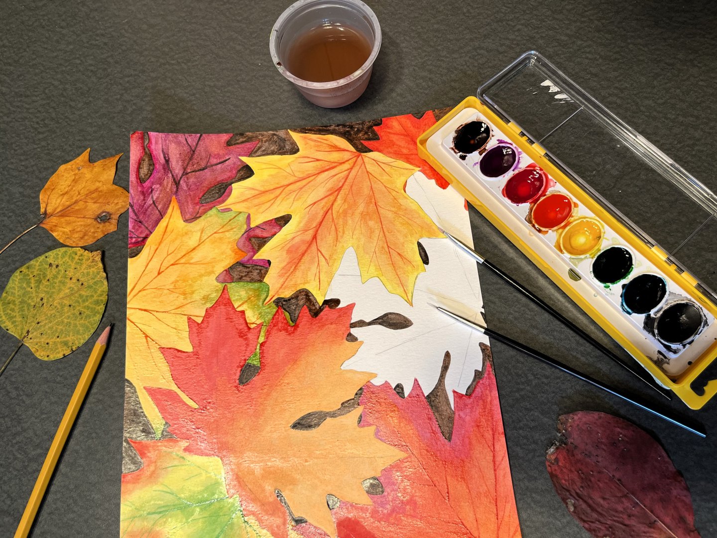 Autumn leaf watercolor painting with brush, palette, pencils, and tea on a dark table
