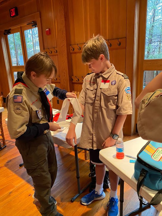 Two Boy Scouts working on a craft at a table in a wood-paneled room.