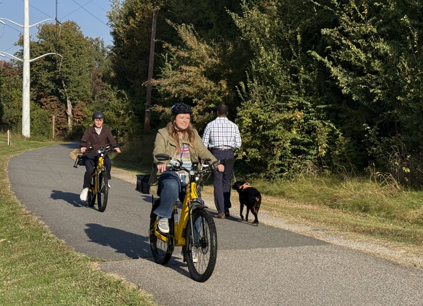 Two cyclists on eBikes and a man walking a dog on a paved path beside trees and grass.